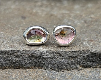 May include: A pair of stud earrings featuring irregular, faceted, multi-colored tourmaline gemstones set in polished silver bezels. The stones display gradients of pink, green, and yellow hues with a translucent, reflective surface texture.