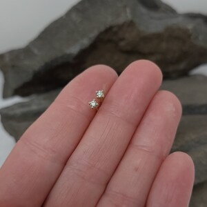 May include: A pair of small, gold-colored stud earrings, each featuring a single, sparkling diamond. The earrings are held between two fingers, showcasing their delicate design and size.