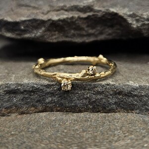 May include: A gold ring with a band textured to resemble a thin tree branch. The ring features two small, round, light brown gemstones set at different points along the branch-like band, creating a delicate and organic appearance.