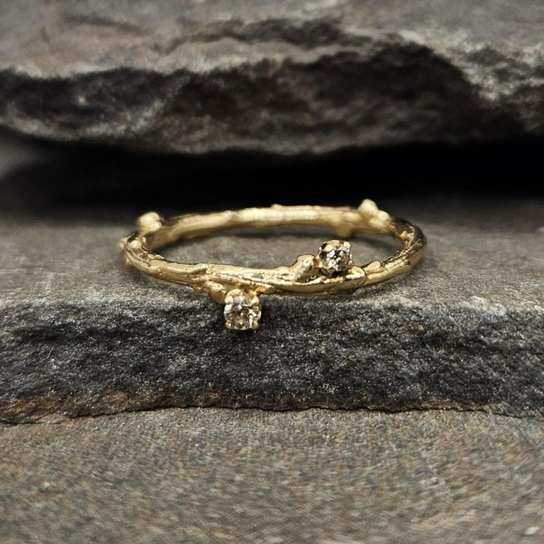 May include: A gold ring with a band textured to resemble a thin tree branch. The ring features two small, round, light brown gemstones set at different points along the branch-like band, creating a delicate and organic appearance.