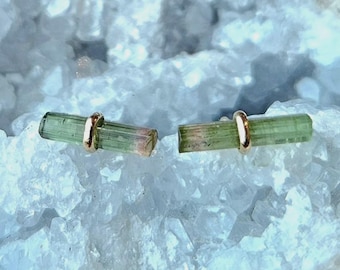 May include: A pair of stud earrings featuring elongated, cylindrical green tourmaline crystals. Each crystal is horizontally oriented and secured in the center by a polished gold-toned metal band, creating a simple, linear design.