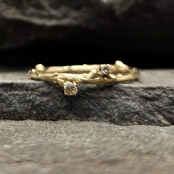 May include: A gold ring with a textured band resembling a small tree branch. The ring features two small, round, clear gemstones set into the metal, positioned at different points along the organic, twig-like structure of the band.