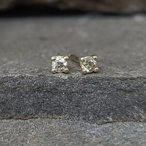 May include: A pair of small, square-shaped stud earrings with a single, round, clear gemstone in each. The earrings are set in a light-colored metal, possibly gold or silver, and are displayed on a textured, gray surface.