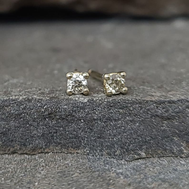May include: A pair of small, square-shaped stud earrings with a single, round, clear gemstone in each. The earrings are set in a light-colored metal, possibly gold or silver, and are displayed on a textured, gray surface.