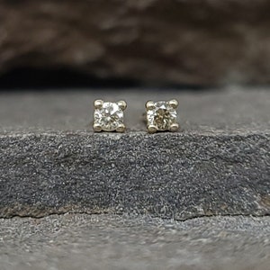 May include: A pair of small, square-shaped stud earrings. Each earring features a round, clear gemstone set in a gold-colored metal. The earrings are displayed on a dark, textured surface.