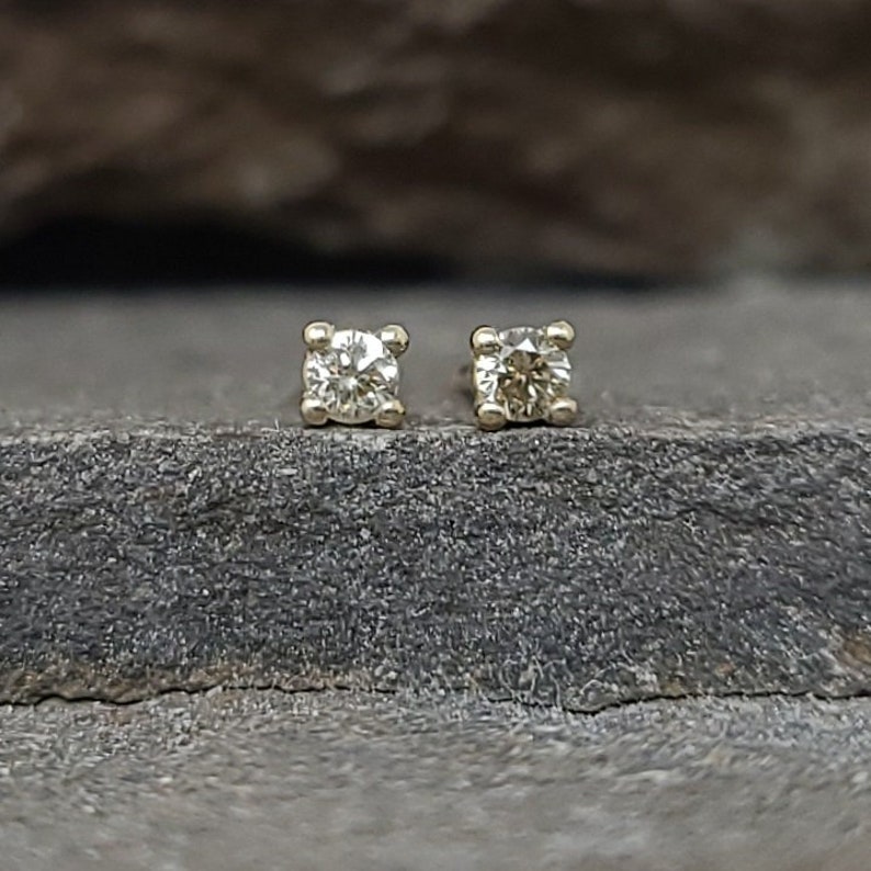 May include: A pair of small, square-shaped stud earrings. Each earring features a round, clear gemstone set in a gold-colored metal. The earrings are displayed on a dark, textured surface.