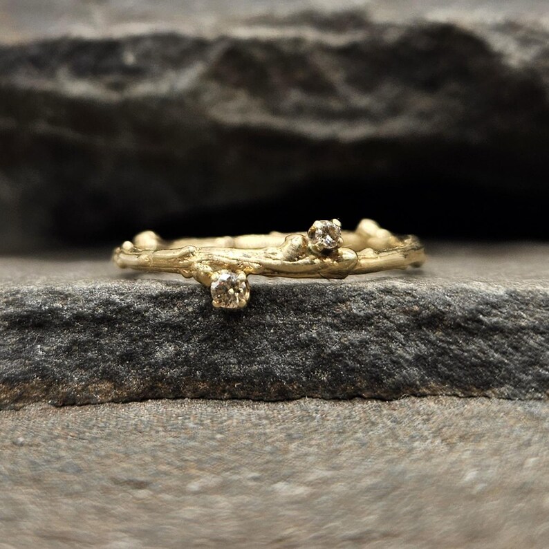 May include: A gold ring with a textured band resembling a natural twig. The ring features two small, round, clear stones set into the metal, mimicking the appearance of buds or small growths on the branch-like structure of the band.