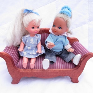 May include: Two dolls, one with blonde hair and a blue dress, the other with blonde hair and a blue shirt and jeans, are sitting on a small wooden sofa with a pink and white striped fabric.