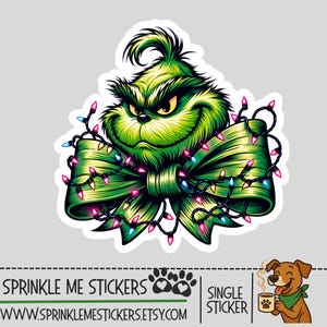 May include: A die-cut sticker featuring a green, furry, scowling character head positioned above a large green ribbon bow wrapped in a string of colorful, glowing Christmas lights. The sticker includes the text SPRINKLE ME STICKERS and a website URL.