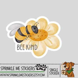 May include: A sticker featuring a cartoon bee with black and yellow stripes, perched on a yellow flower. The text "BEE KIND" is below the bee. The sticker has a white border and is on a gray background.