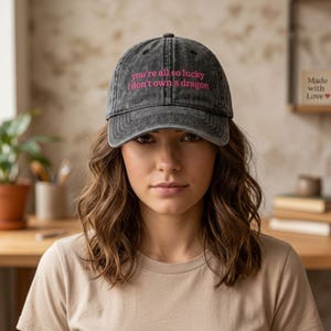 Könnte beinhalten: Dunkelgraue Baseballkappe mit dem Text "you're all so lucky I don't own a dragon" in Pink. Die Kappe wird von einer Person getragen, die ein beiges T-Shirt trägt.