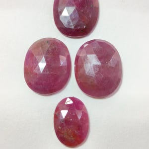 May include: Four faceted oval-shaped rubies in varying sizes. The gemstones are a deep pinkish-red colour, with visible internal inclusions and a polished, reflective surface. The gems are arranged on a white background.