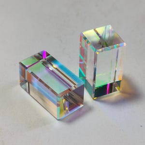 May include: Two rectangular glass prisms with iridescent color reflections. One prism is lying down, and the other is standing upright. The prisms are clear and have a polished appearance, reflecting light in a rainbow of colors.