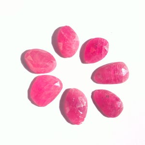Può includere: Sette gemme rosa ovali e sfaccettate, disposte a cerchio. Le gemme sono molto lucide e riflettono la luce, proiettando ombre sullo sfondo bianco. Le gemme sono di un rosa vibrante.