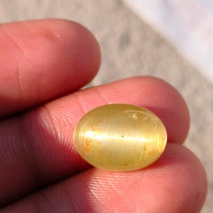 Puede incluir: Una gema de color amarillo dorado con forma ovalada y redondeada. La superficie pulida refleja la luz, creando una banda horizontal brillante en el centro. La gema se sostiene entre dos dedos, mostrando su color y claridad.