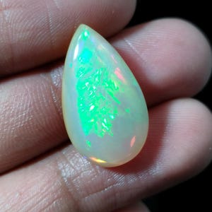 May include: A pear-shaped opal gemstone with a creamy white base color. The stone displays vibrant flashes of green and other colors, creating a play-of-color effect. The polished surface reflects light, highlighting the internal patterns.