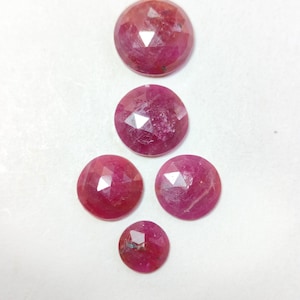 May include: Five faceted, round-cut rubies in varying sizes are arranged on a white surface. The gemstones display a deep red hue with internal variations, reflecting light across their cut surfaces. The arrangement showcases the gems' polished facets.
