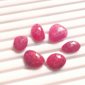 May include: Seven faceted, oval-shaped, and irregular-shaped ruby gemstones. The rubies are a deep, vibrant pinkish-red color. The gemstones are displayed on a white, ridged surface, creating a contrast that highlights their color and shape.