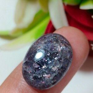 Puede incluir: Una gema pulida de forma ovalada, con un color base p&uacute;rpura oscuro y motas iridiscentes rosas, azules y plateadas. La piedra se sostiene entre dos dedos, mostrando su superficie lisa y reflectante. La textura de la gema es cristalina.