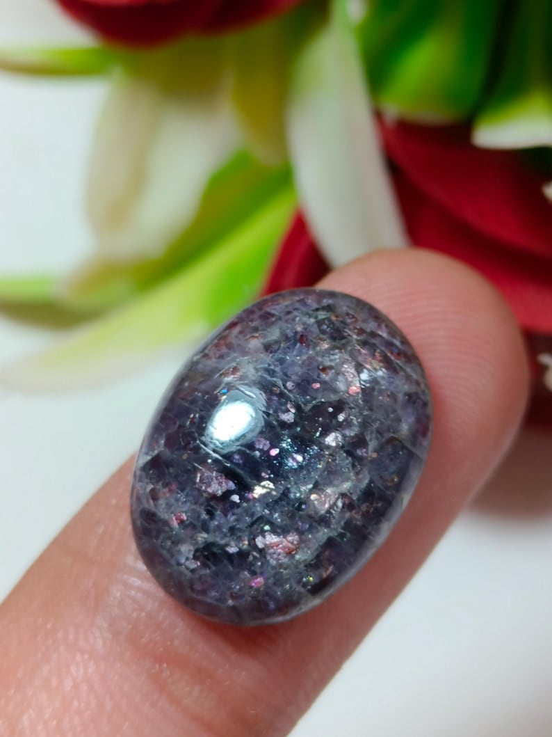 Puede incluir: Una gema pulida de forma ovalada, con un color base p&uacute;rpura oscuro y motas iridiscentes rosas, azules y plateadas. La piedra se sostiene entre dos dedos, mostrando su superficie lisa y reflectante. La textura de la gema es cristalina.