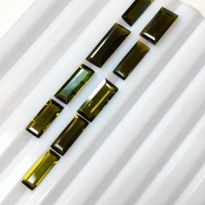 May include: A collection of eight rectangular baguette-cut gemstones in varying shades of olive green. The stones are arranged in two parallel rows, each featuring a faceted surface that reflects light, showcasing deep forest and lime green tones.