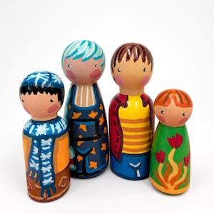 Peut inclure: Quatre poupées en bois peintes à la main disposées en rangée. La première porte un bonnet et une écharpe bleus sur un manteau orange. La deuxième a des cheveux bleu sarcelle et un tablier bleu foncé à étoiles orange. La troisième porte un haut jaune rayé et une veste rouge. La quatrième est verte avec des tulipes rouges.