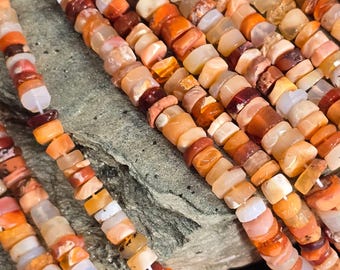 May include: Close-up of several strands of gemstone beads in shades of orange, red, and cream. The beads are small, irregular shapes, strung together to create a necklace or bracelet. The background is a textured, gray surface.