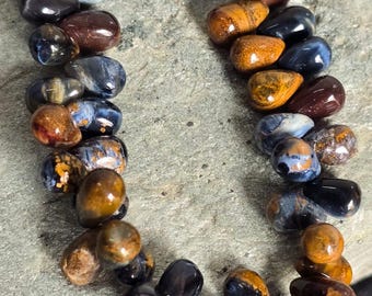 May include: A close-up of a necklace made of polished, teardrop-shaped beads. The beads are a mix of colors, including blue, brown, and gold. The necklace is resting on a gray, textured surface.