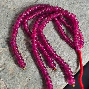 Puede incluir: Primer plano de varios hilos de pequeñas cuentas redondas de color rosa intenso. Las cuentas están ensartadas juntas, con una cuerda roja visible en el extremo. Las cuentas tienen un acabado brillante y están dispuestas sobre una superficie gris texturizada.