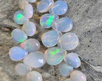 May include: Strands of opalescent, teardrop-shaped opal beads. The beads are strung together, displaying a range of iridescent colors, including blues, greens, and pinks. The opals have a smooth, polished surface, reflecting light and creating a shimmering effect.