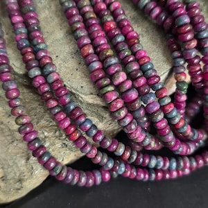 Może przedstawiać: Zbliżenie na wiele sznurów koralików z kamieni szlachetnych. Koraliki są w odcieniach głębokiej czerwieni, fioletu i niebieskiego. Małe, okrągłe koraliki są nawleczone, tworząc teksturowany efekt. Idealne do tworzenia biżuterii.