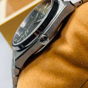 May include: A silver-toned stainless steel wristwatch featuring a dark green textured dial with a waffle pattern, a black bezel, and a metallic link bracelet. The side of the case displays a crown embossed with a T logo, showcasing a sleek design.