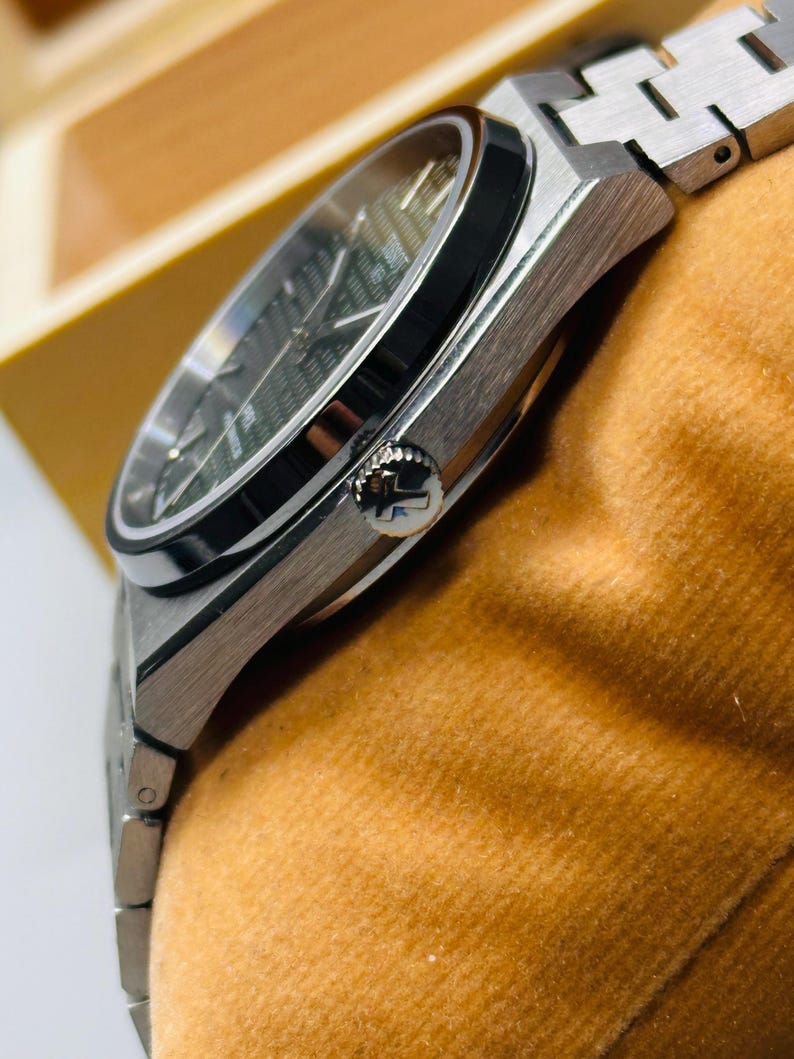 May include: A silver-toned stainless steel wristwatch featuring a dark green textured dial with a waffle pattern, a black bezel, and a metallic link bracelet. The side of the case displays a crown embossed with a T logo, showcasing a sleek design.