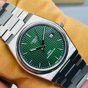 May include: A Tissot PRX Powermatic 80 wristwatch with a brushed stainless steel integrated bracelet and a round case. The dial is a deep green with a waffle-patterned texture, silver-tone hands, index markers, and a date window at the 3 o'clock position.
