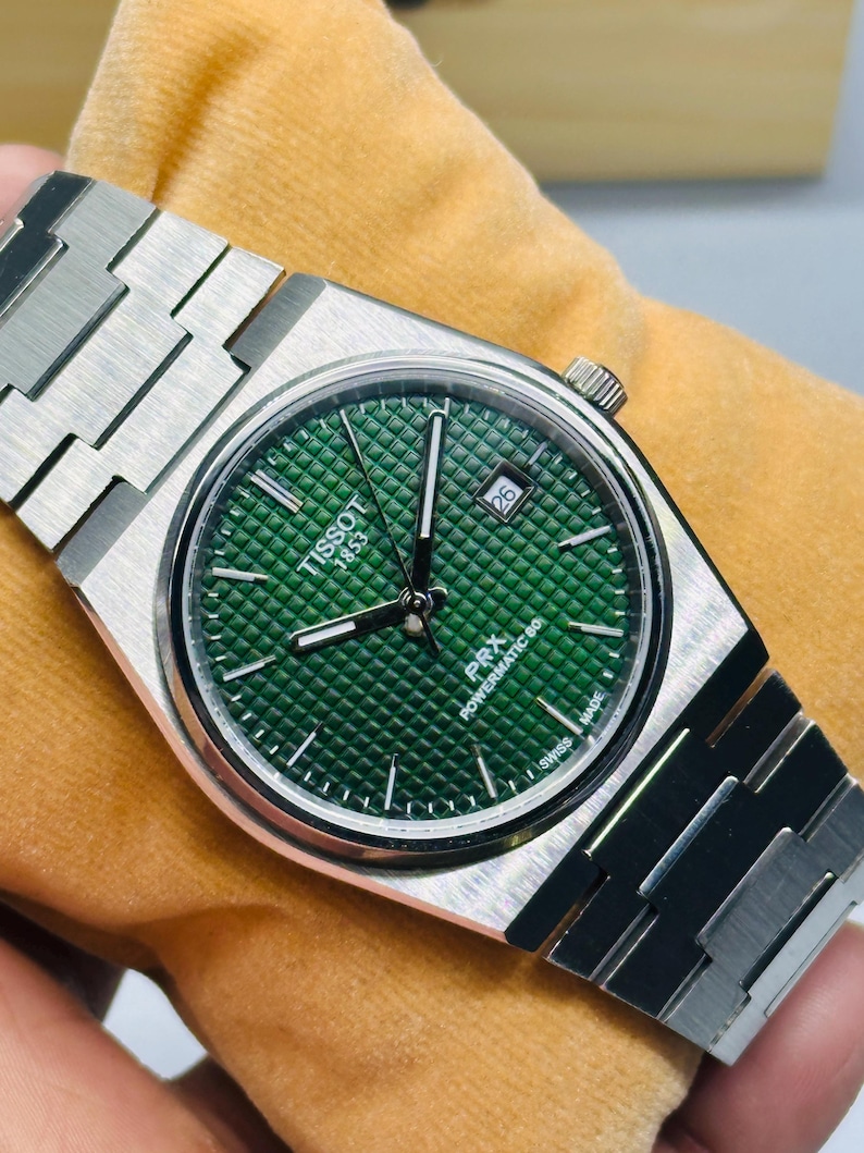 May include: A Tissot PRX Powermatic 80 wristwatch with a brushed stainless steel integrated bracelet and a round case. The dial is a deep green with a waffle-patterned texture, silver-tone hands, index markers, and a date window at the 3 o'clock position.