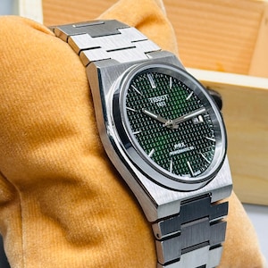 May include: A Tissot 1853 PRX Powermatic 80 wristwatch featuring a brushed stainless steel case and integrated bracelet. The dial is a deep green with a waffle-patterned texture, silver-toned hands, and a date window at the three o'clock position.