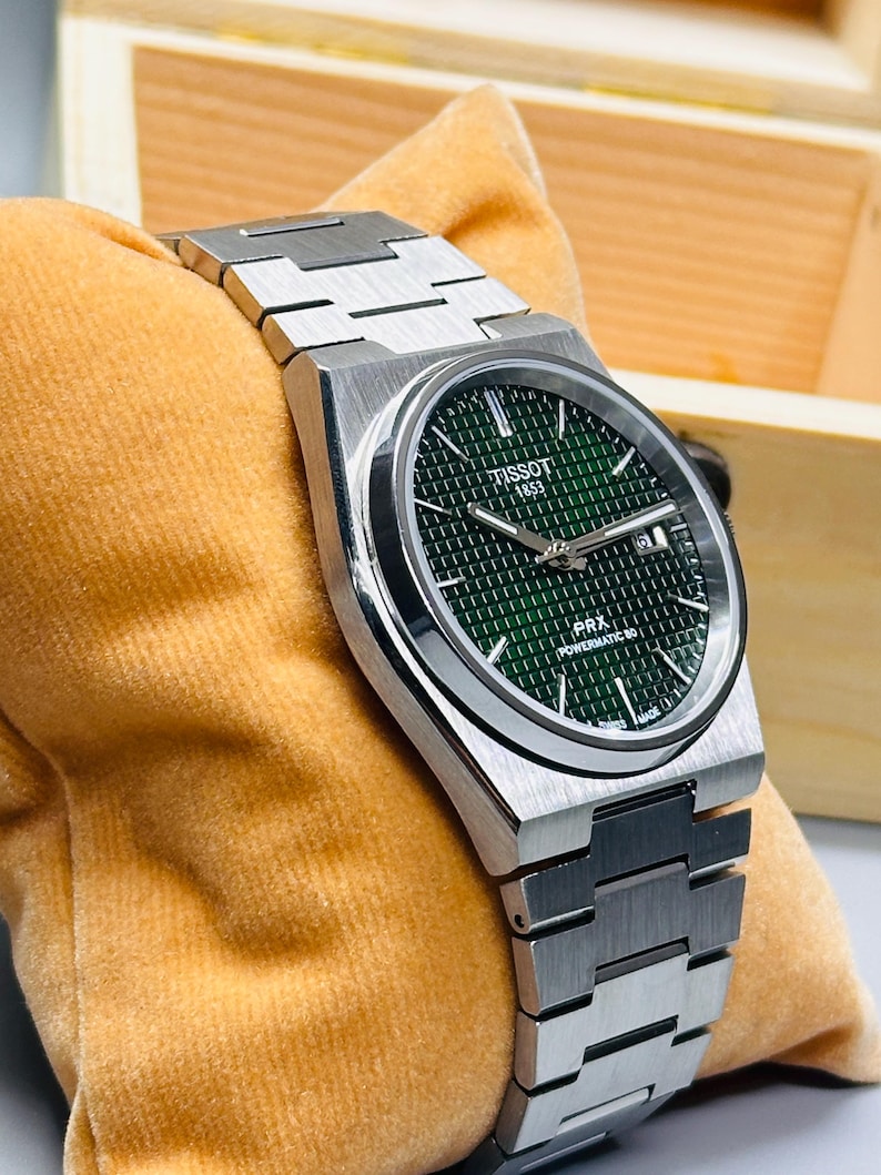 May include: A Tissot 1853 PRX Powermatic 80 wristwatch featuring a brushed stainless steel case and integrated bracelet. The dial is a deep green with a waffle-patterned texture, silver-toned hands, and a date window at the three o'clock position.