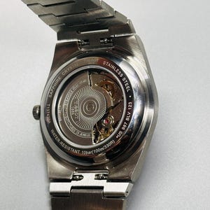May include: A silver-toned stainless steel Tissot wristwatch with an exhibition case back revealing the internal movement. The back is engraved with Tissot 1853, Sapphire Crystal, Stainless Steel, Water-Resistant 10bar (100m/330ft), and T137423A.