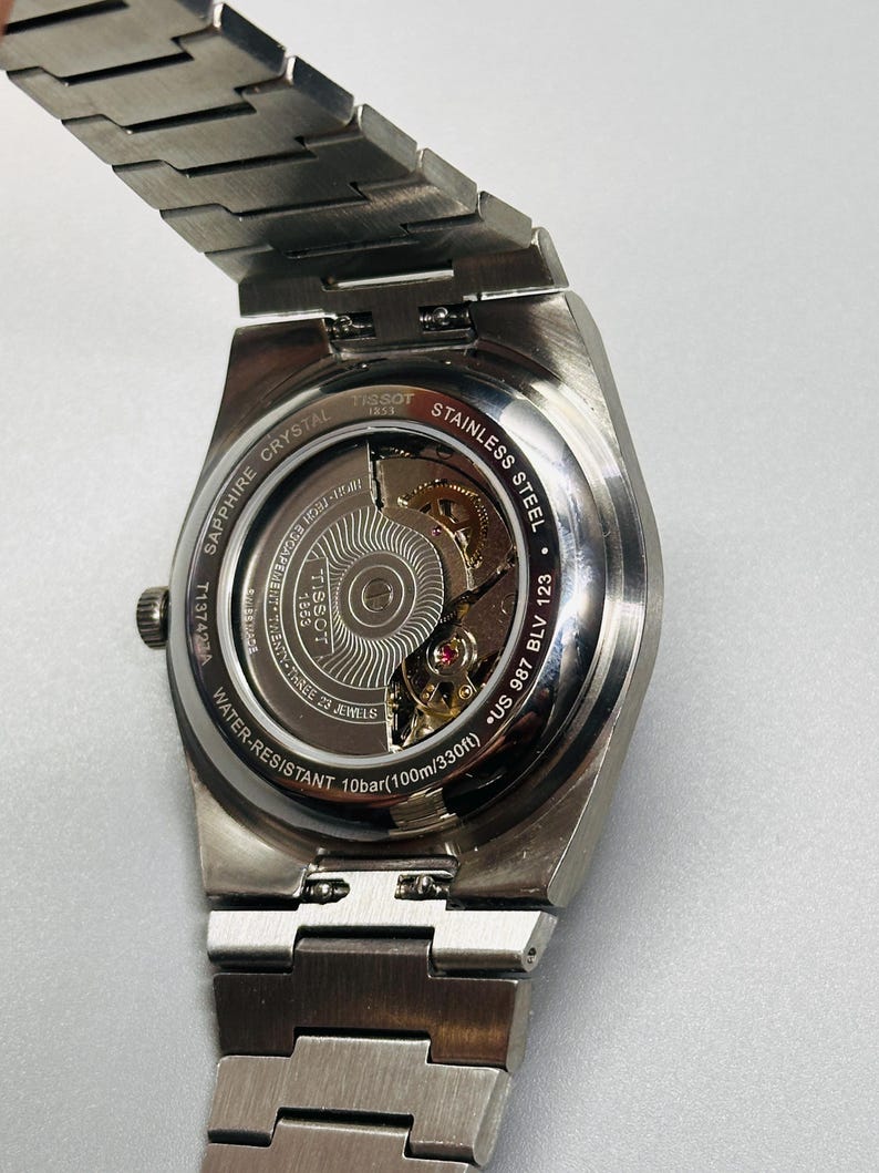 May include: A silver-toned stainless steel Tissot wristwatch with an exhibition case back revealing the internal movement. The back is engraved with Tissot 1853, Sapphire Crystal, Stainless Steel, Water-Resistant 10bar (100m/330ft), and T137423A.