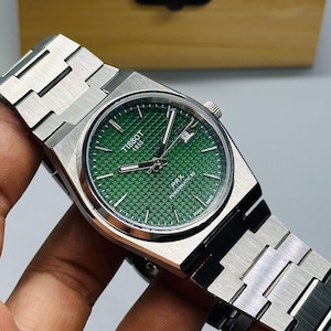 May include: A Tissot PRX Powermatic 80 wristwatch with a green waffle-patterned dial, silver-tone hands, and a date window at the three o'clock position. The watch features a brushed stainless steel case and an integrated link bracelet.