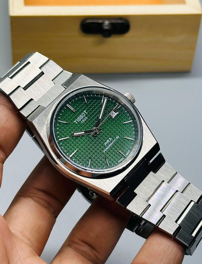 May include: A Tissot PRX Powermatic 80 wristwatch with a green waffle-patterned dial, silver-tone hands, and a date window at the three o'clock position. The watch features a brushed stainless steel case and an integrated link bracelet.