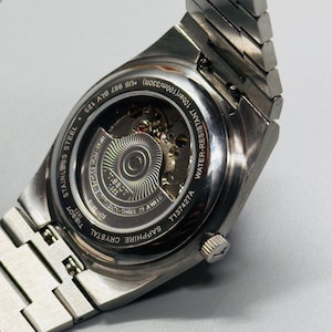 May include: A silver-toned stainless steel wristwatch with an exhibition case back revealing the internal mechanical movement. The watch features a metal link bracelet, a sapphire crystal back, and is water-resistant up to 100m/330ft.