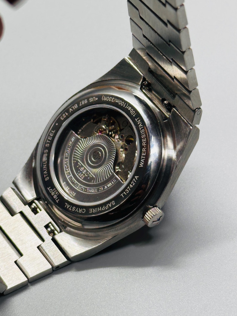 May include: A silver-toned stainless steel wristwatch with an exhibition case back revealing the internal mechanical movement. The watch features a metal link bracelet, a sapphire crystal back, and is water-resistant up to 100m/330ft.