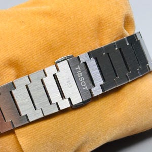 May include: A silver-toned stainless steel watch bracelet featuring a brushed metal finish. The clasp is engraved with the text TISSOT and 1853. The links are rectangular and connected in a standard link design, showcasing a metallic sheen.
