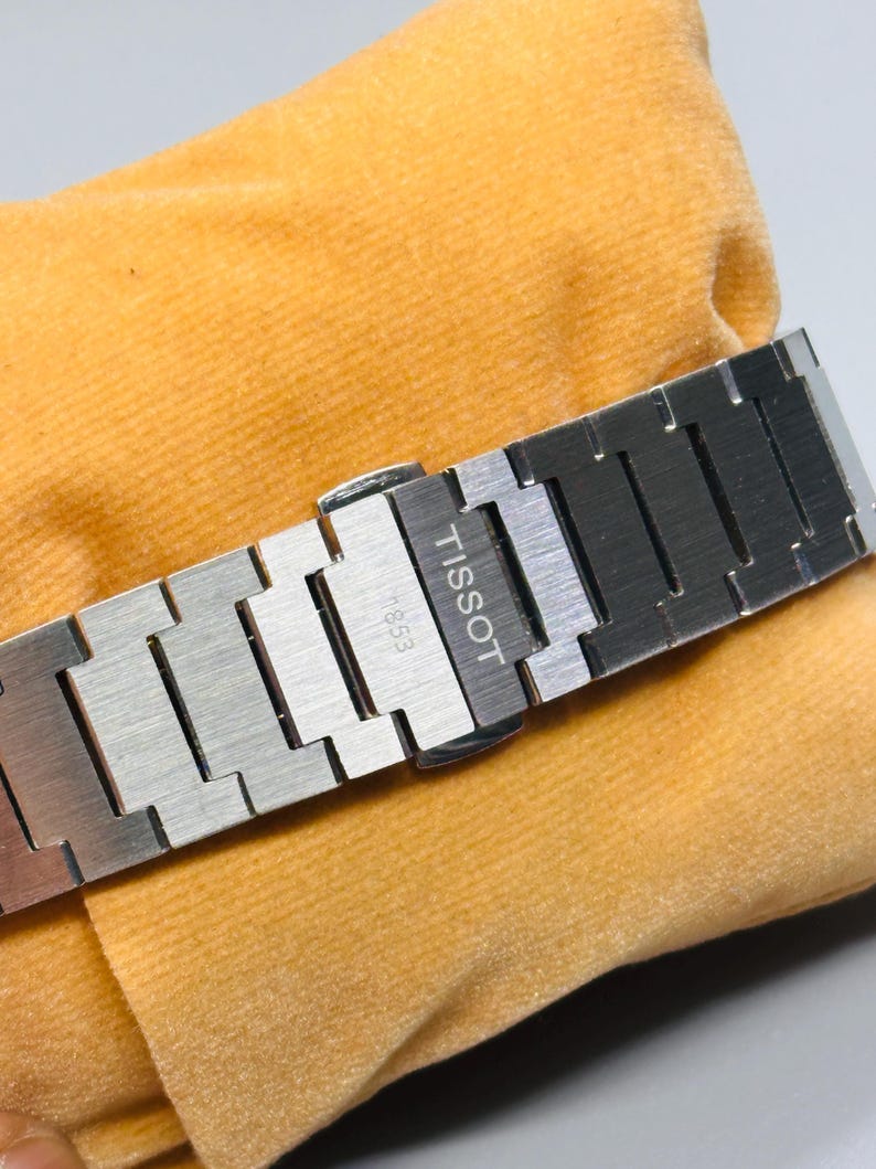 May include: A silver-toned stainless steel watch bracelet featuring a brushed metal finish. The clasp is engraved with the text TISSOT and 1853. The links are rectangular and connected in a standard link design, showcasing a metallic sheen.