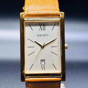 May include: A rectangular Seiko watch with a gold-tone case and a light-coloured face. The watch features Roman numeral hour markers, a date display, and gold hands. The watch has a brown leather strap.