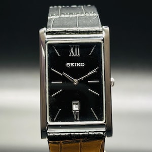 May include: A rectangular Seiko watch with a black face, silver case, and a leather strap. The watch face features silver hands and markers, with the number XII at the top and VI at the bottom. The date is displayed at the 6 o'clock position.