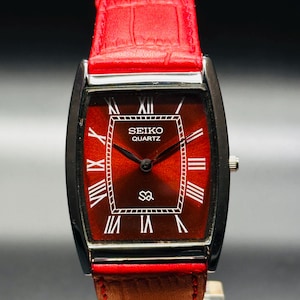 May include: A rectangular Seiko Quartz watch with a red face and black frame. The watch features white Roman numerals and hands, with a red leather strap. The watch face displays the Seiko Quartz logo.