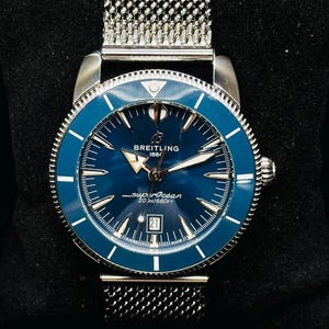 May include: A Breitling Superocean watch with a blue dial and bezel, silver-tone hands and indices, and a stainless steel mesh bracelet. The watch face displays the text Breitling 1884, superocean, 200M/660FT, and a date window showing the number 21.