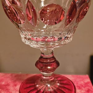 Puede incluir: Una copa de champán de cristal transparente con base y detalles en rojo rubí. El cuenco presenta un patrón tallado con diseños ovalados y circulares. El tallo tiene forma bulbosa y la base un diseño de estrella.